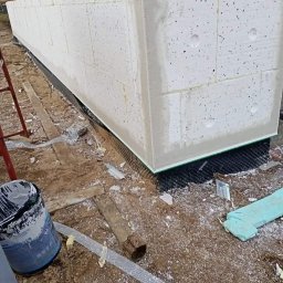 Usługi remontowo - budowlane REM-BUD - Narożnik budynku w trakcie ocieplania, widoczne białe bloki styropianowe, siatka zbrojeniowa, grunt pod ocieplenie oraz fragment drabiny malarskiej.