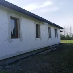 Ściana budynku z betonu komórkowego po wstępnym ociepleniu, z widocznymi fragmentami szpachli wokół okien i niedoskonałościami powierzchni, na tle jesiennego krajobrazu.