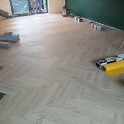 Usługi remontowo - budowlane REM-BUD - Układanie paneli podłogowych w jodełkę, widoczny fragment podkładu, paczki paneli, rolka żółtej folii i drabina oparta o zieloną ścianę w tle.