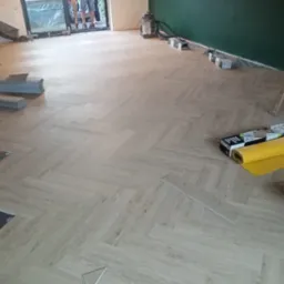 Układanie paneli podłogowych w jodełkę, widoczny fragment podkładu, paczki paneli, rolka żółtej folii i drabina oparta o zieloną ścianę w tle.