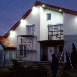 Usługi remontowo - budowlane REM-BUD - Dwupiętrowy dom jednorodzinny w trakcie prac elewacyjnych, oświetlony reflektorami po zmroku, z widocznymi rusztowaniami i częściowo pomalowaną fasadą.