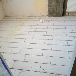 Usługi remontowo - budowlane REM-BUD - Ułożona jasna podłoga z płytek imitujących drewno w trakcie remontu, widoczne krzyżyki dystansowe, ściany częściowo przygotowane pod malowanie, grzejnik.