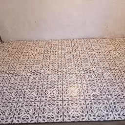 Usługi remontowo - budowlane REM-BUD - Ułożona podłoga z białych płytek z ciemnym, wzorzystym ornamentem w pomieszczeniu z surowymi, szarymi ścianami i widoczną instalacją elektryczną w rogu.