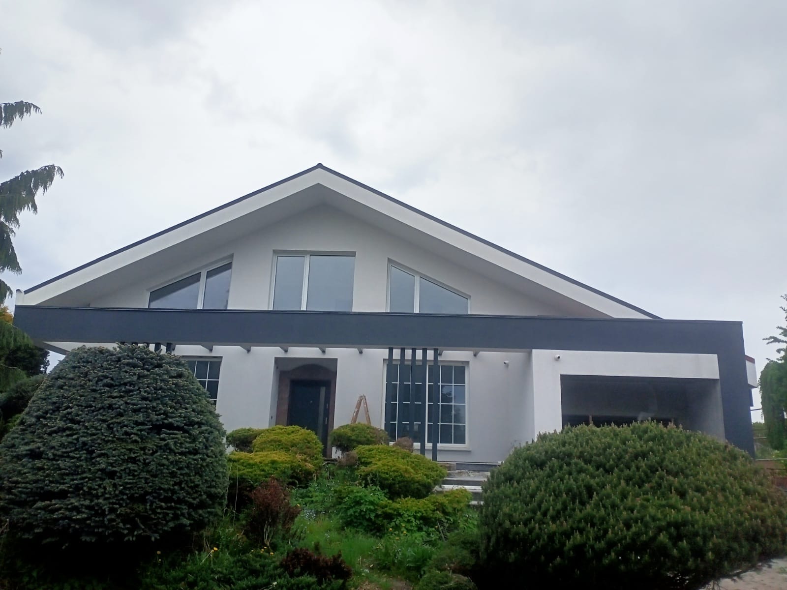 Nowoczesny dom jednorodzinny z białą elewacją, grafitowymi akcentami i dużymi oknami, otoczony bujną zielenią w pochmurny dzień.