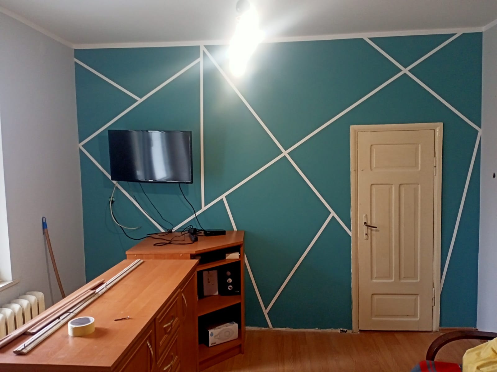 Pomieszczenie po malowaniu ścian na kolor morski z białymi geometrycznymi wzorami, widoczny telewizor, meble i drzwi