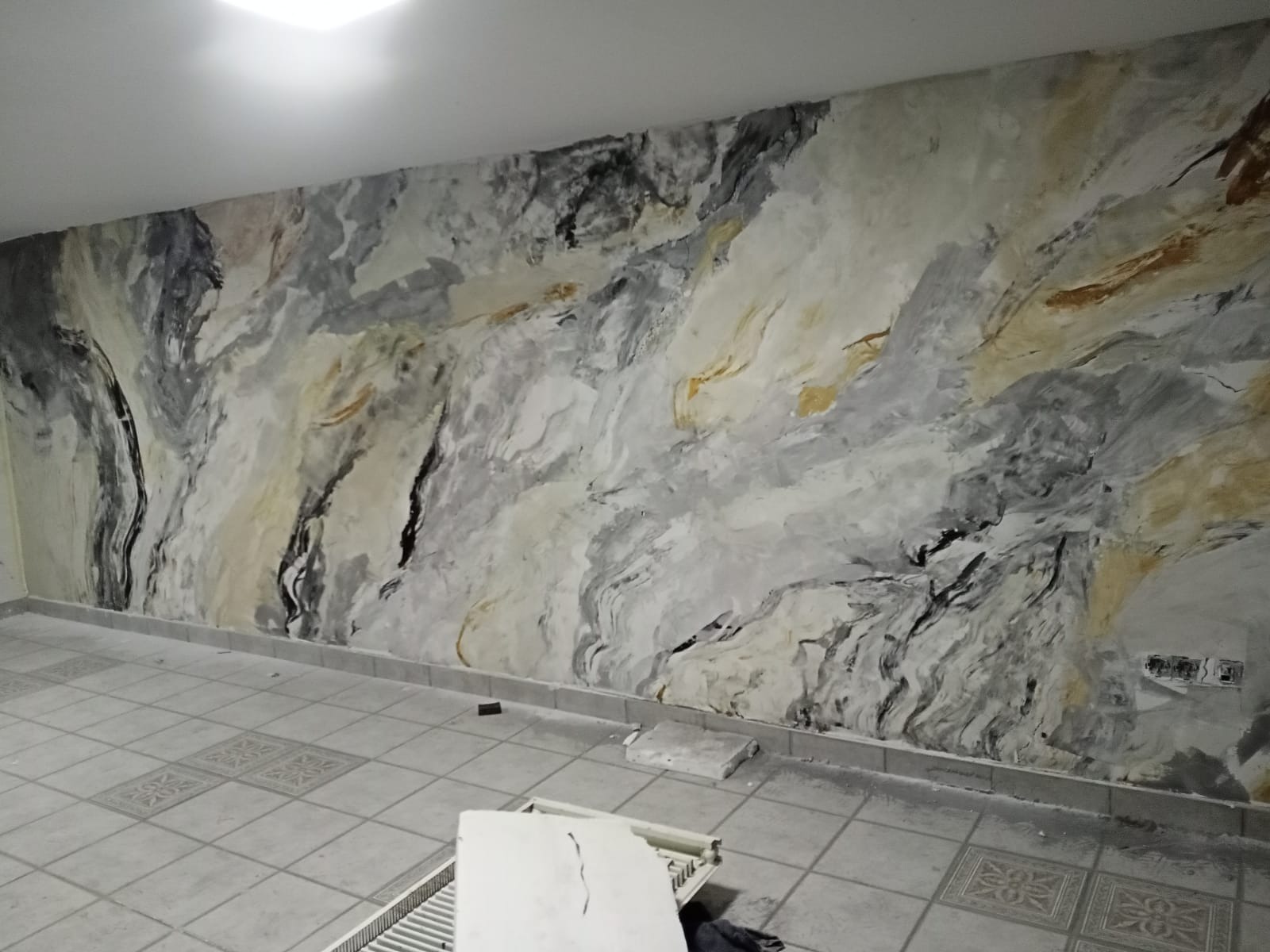 Ściana w pokoju wykończona dekoracyjnym efektem imitującym marmur w odcieniach szarości, bieli, czerni i złota, widoczne płytki podłogowe i fragment grzejnika.