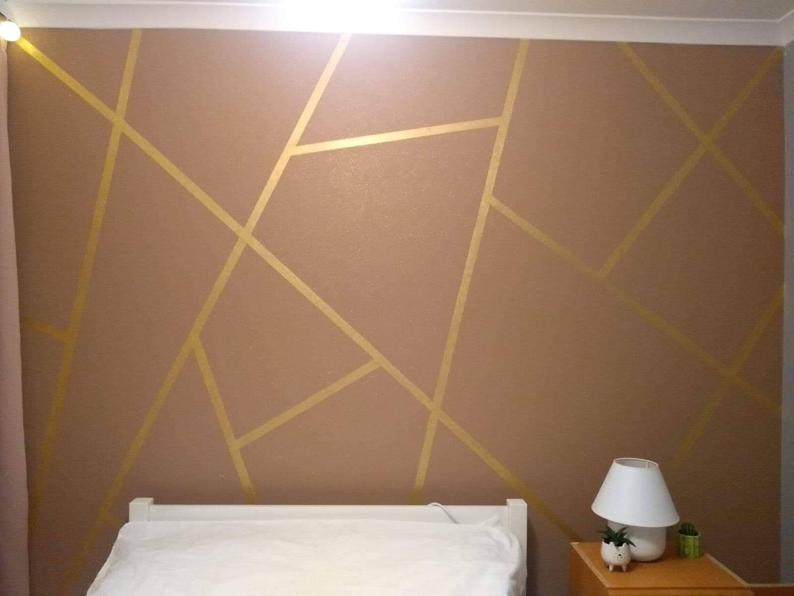 Ściana w sypialni pomalowana na brązowo z geometrycznymi złotymi paskami, białe łóżko z pościelą i lampka nocna z sukulentami.