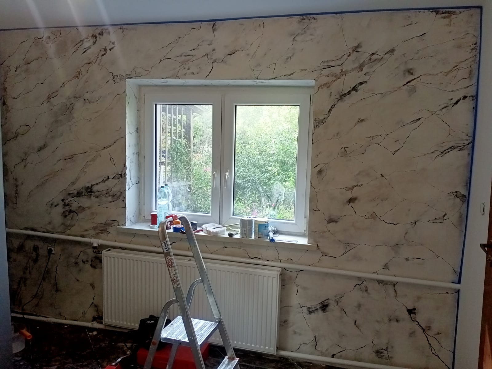 Ściana po dekoracyjnym malowaniu imitującym marmur, zabezpieczona taśmą malarską przy suficie, z grzejnikiem i drabiną w tle, widok na okno z zielenią za nim.