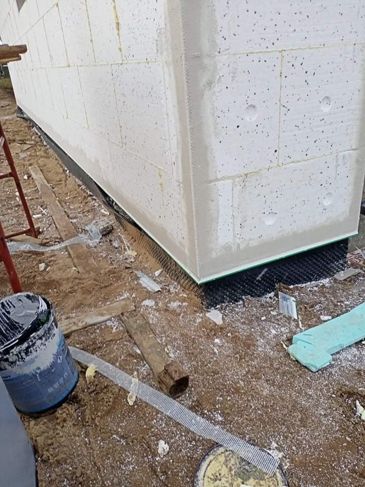 Narożnik budynku w trakcie ocieplania, widoczne białe bloki styropianowe, siatka zbrojeniowa, grunt pod ocieplenie oraz fragment drabiny malarskiej.