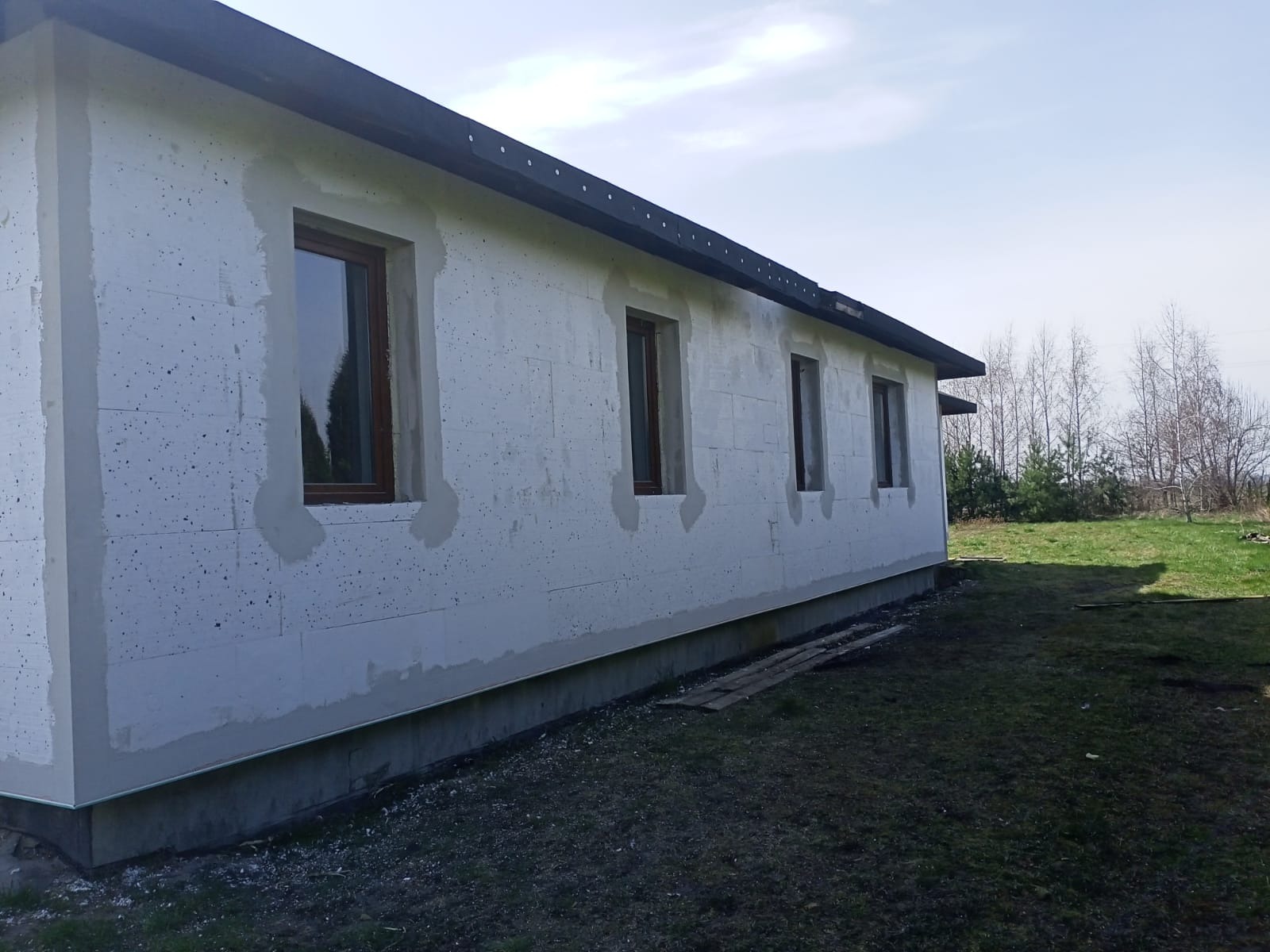 Ściana budynku z betonu komórkowego po wstępnym ociepleniu, z widocznymi fragmentami szpachli wokół okien i niedoskonałościami powierzchni, na tle jesiennego krajobrazu.