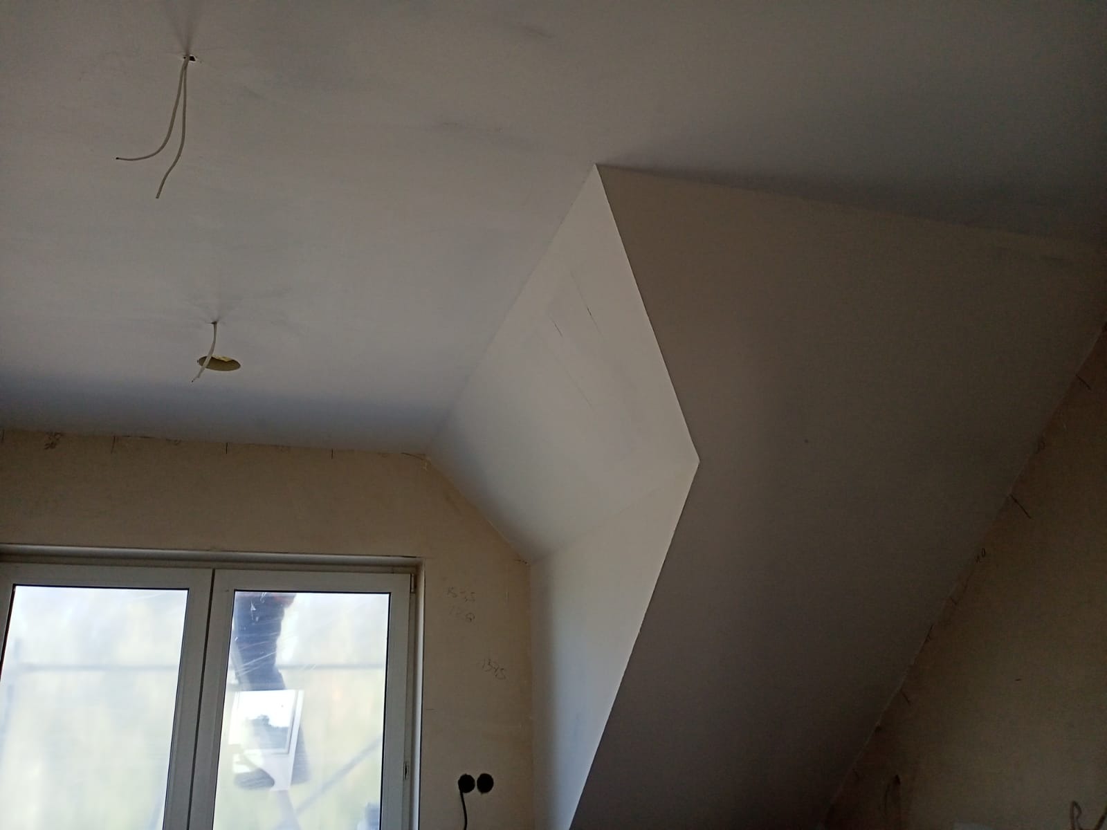 Poddasze po szpachlowaniu z widocznymi przewodami elektrycznymi, okno zasłonięte folią malarską, ściany przygotowane do dalszych prac wykończeniowych.