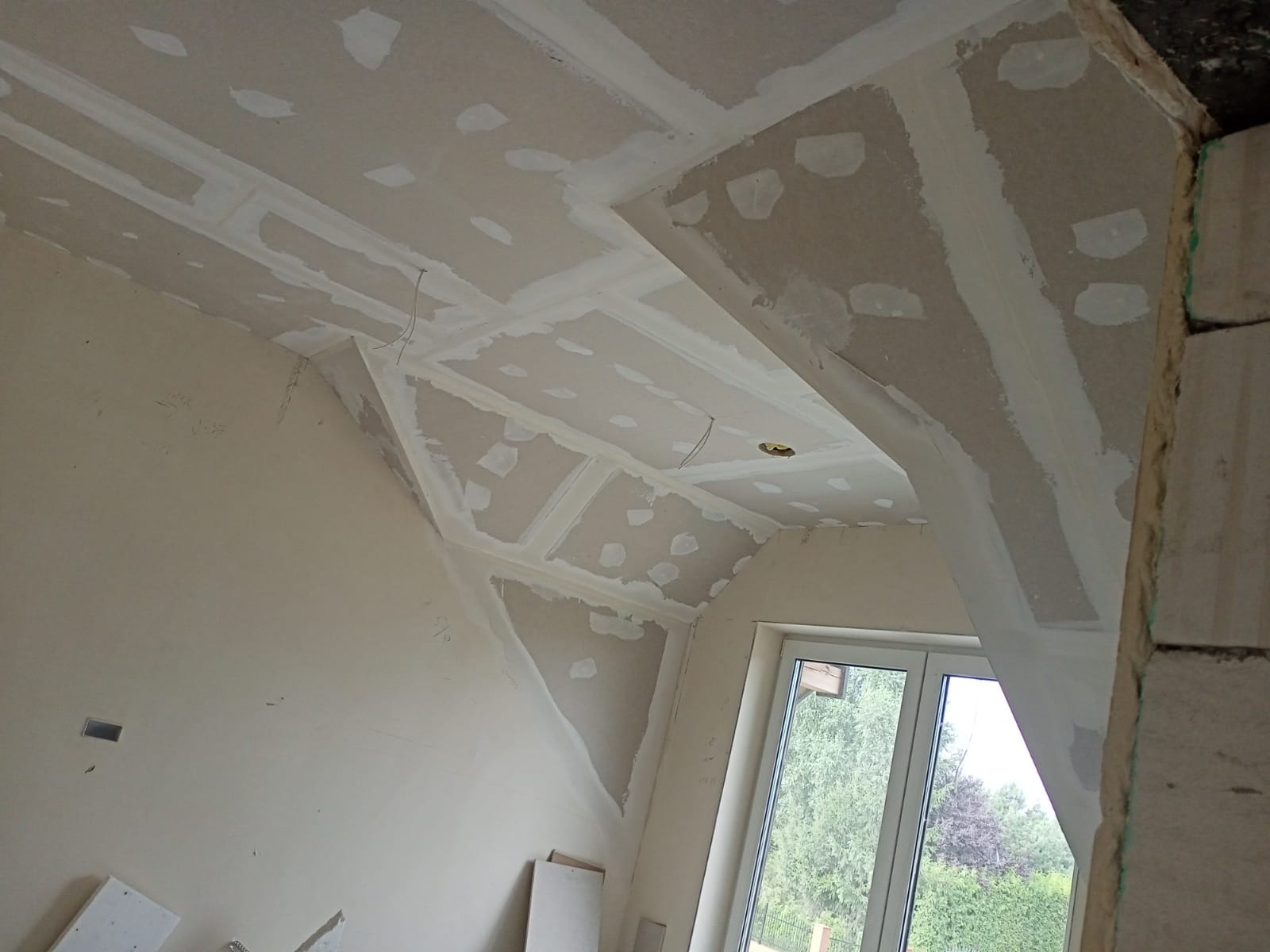 Skośny sufit poddasza z płyt gipsowo-kartonowych w trakcie wykańczania, widoczne łączenia płyt wypełnione masą szpachlową, przygotowane do dalszej obróbki, okno dachowe w tle.