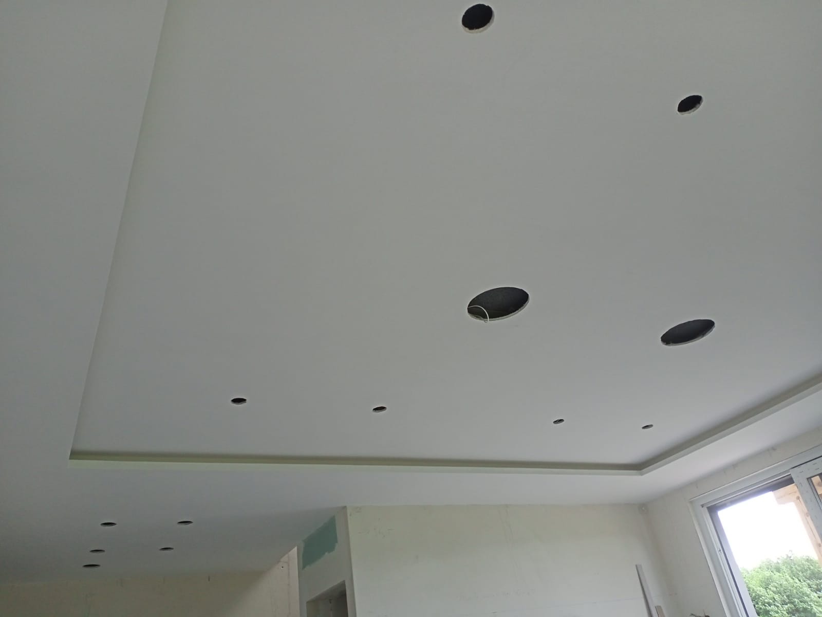 Biały sufit podwieszany z otworami na oświetlenie punktowe, widoczne przewody, ściany w trakcie wykończenia, okno z widokiem na zieleń.