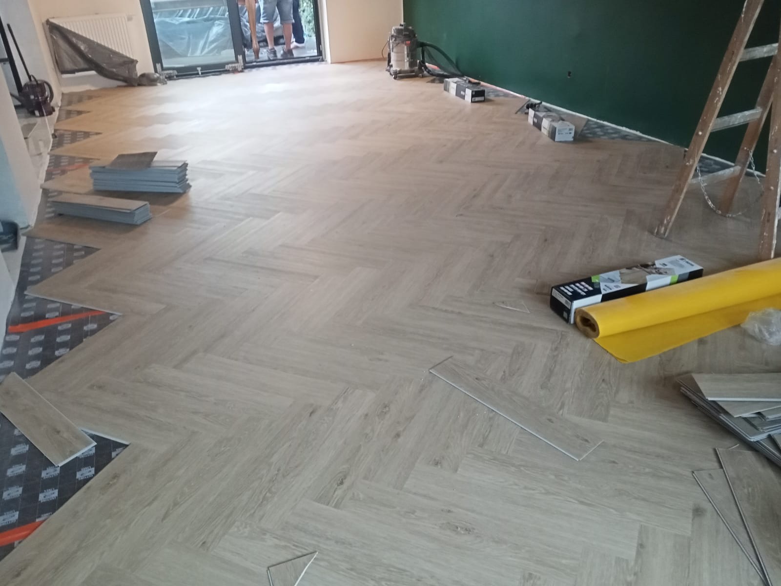 Układanie paneli podłogowych w jodełkę, widoczny fragment podkładu, paczki paneli, rolka żółtej folii i drabina oparta o zieloną ścianę w tle.