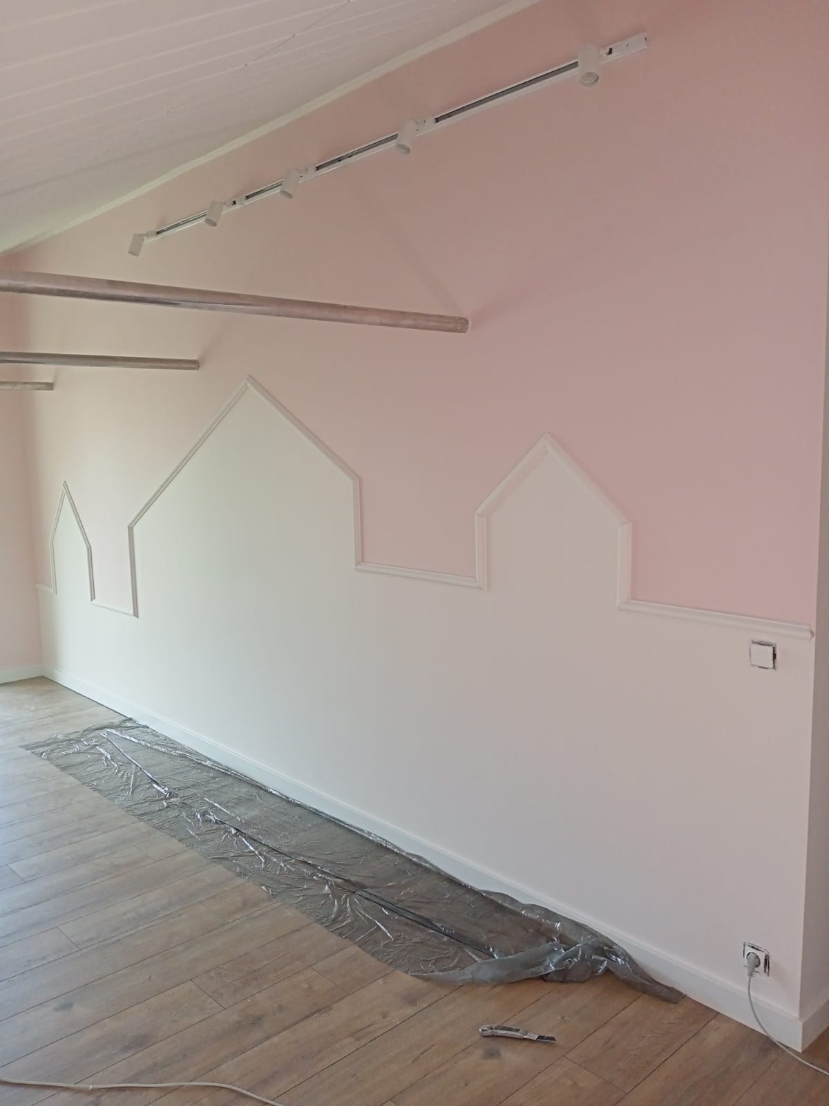 Pokój dziecięcy w trakcie malowania: ściana pomalowana na różowo z białymi obrysami domków, podłoga zabezpieczona folią, widoczne listwy oświetleniowe na suficie.