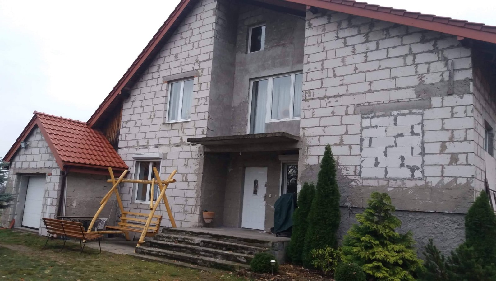 Dom w trakcie ocieplania elewacji, widoczne bloki z betonu komórkowego, częściowo zacierane zaprawą, z elementami przygotowanymi pod okna i drzwi, oraz drewniana huśtawka na pierwszym planie.