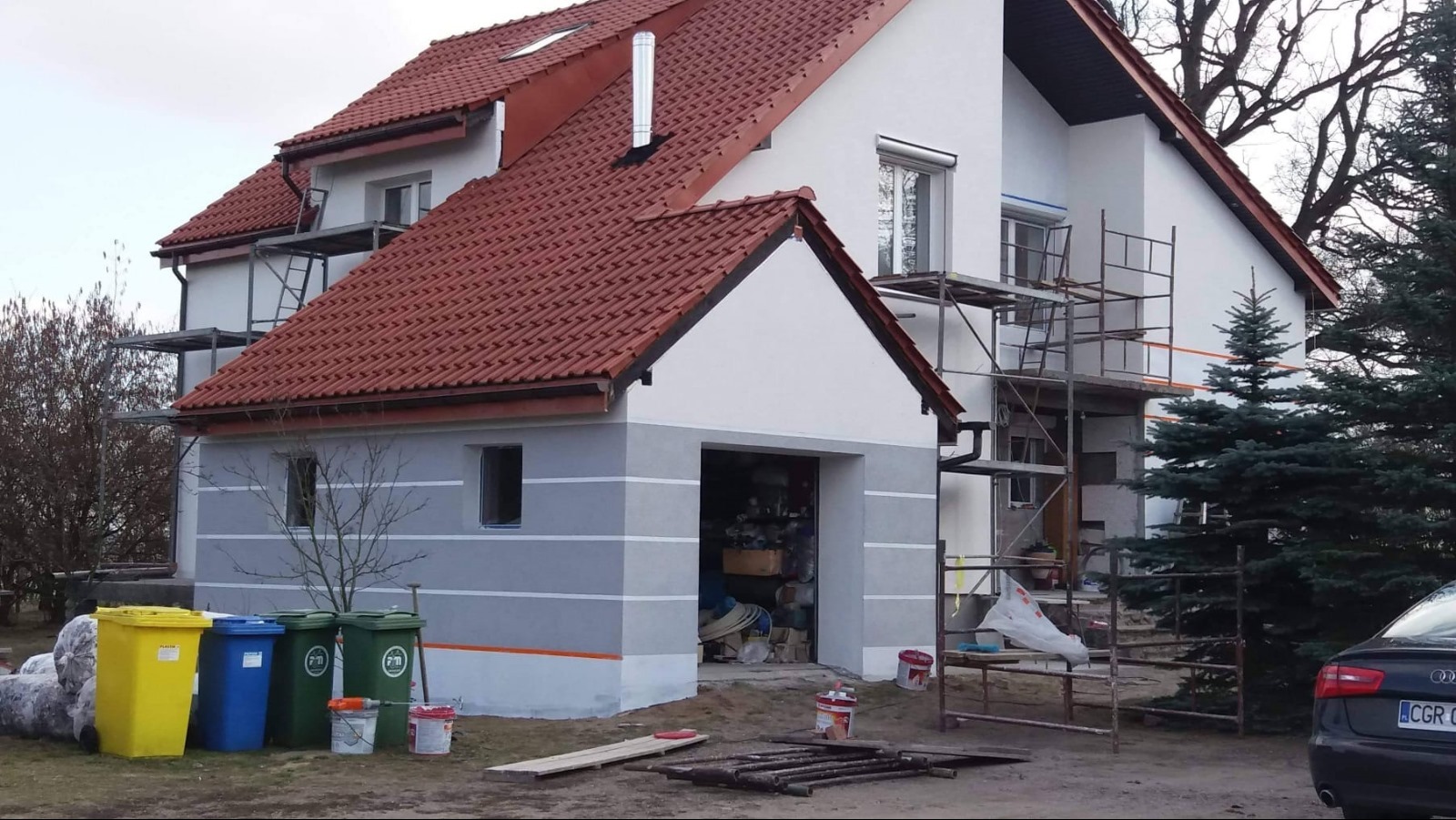 Dwukondygnacyjny dom jednorodzinny w trakcie prac elewacyjnych, z widocznymi rusztowaniami, świeżo pomalowanymi fragmentami ścian na biało i szaro, czerwonym dachem z dachówki, oraz pojemnikami...