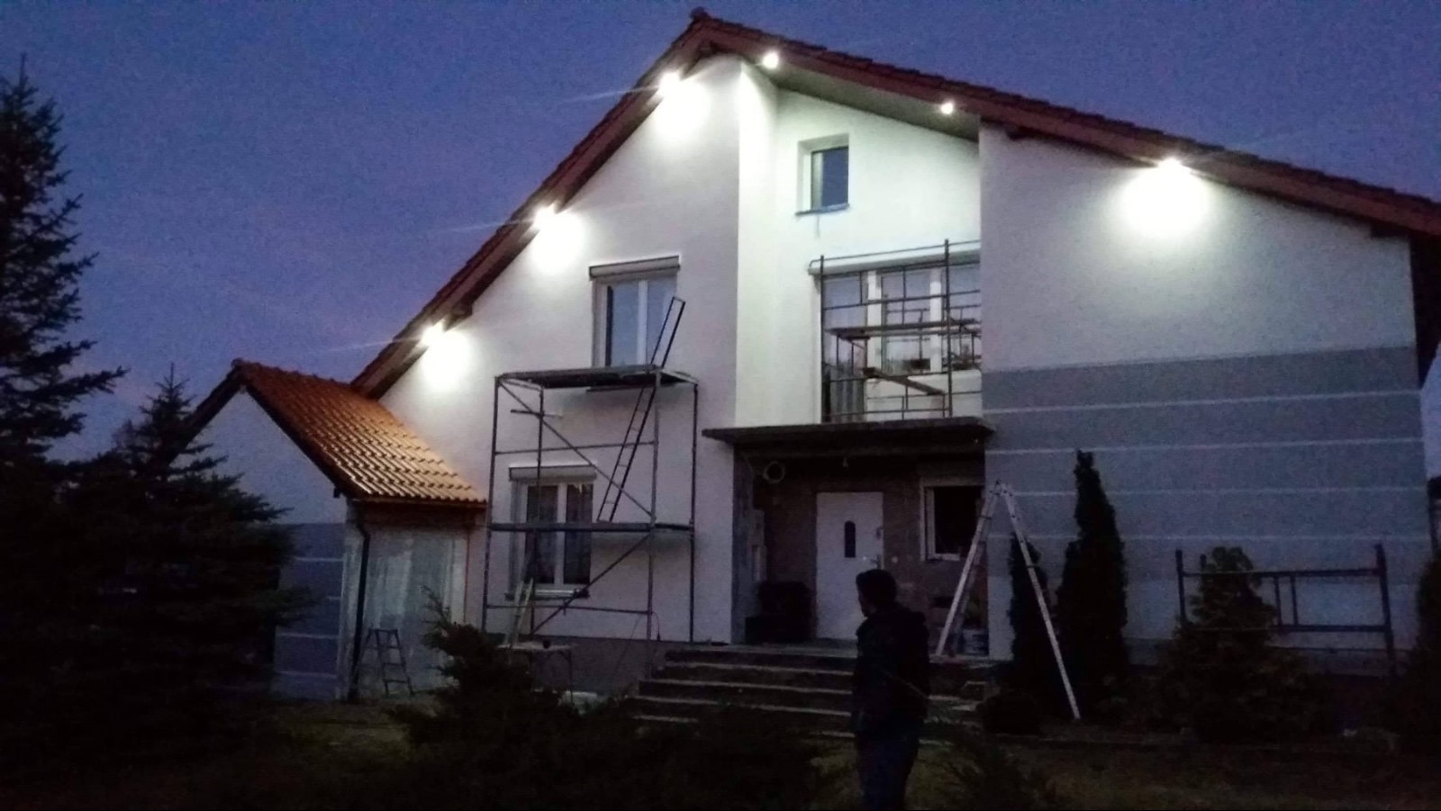 Dwupiętrowy dom jednorodzinny w trakcie prac elewacyjnych, oświetlony reflektorami po zmroku, z widocznymi rusztowaniami i częściowo pomalowaną fasadą.