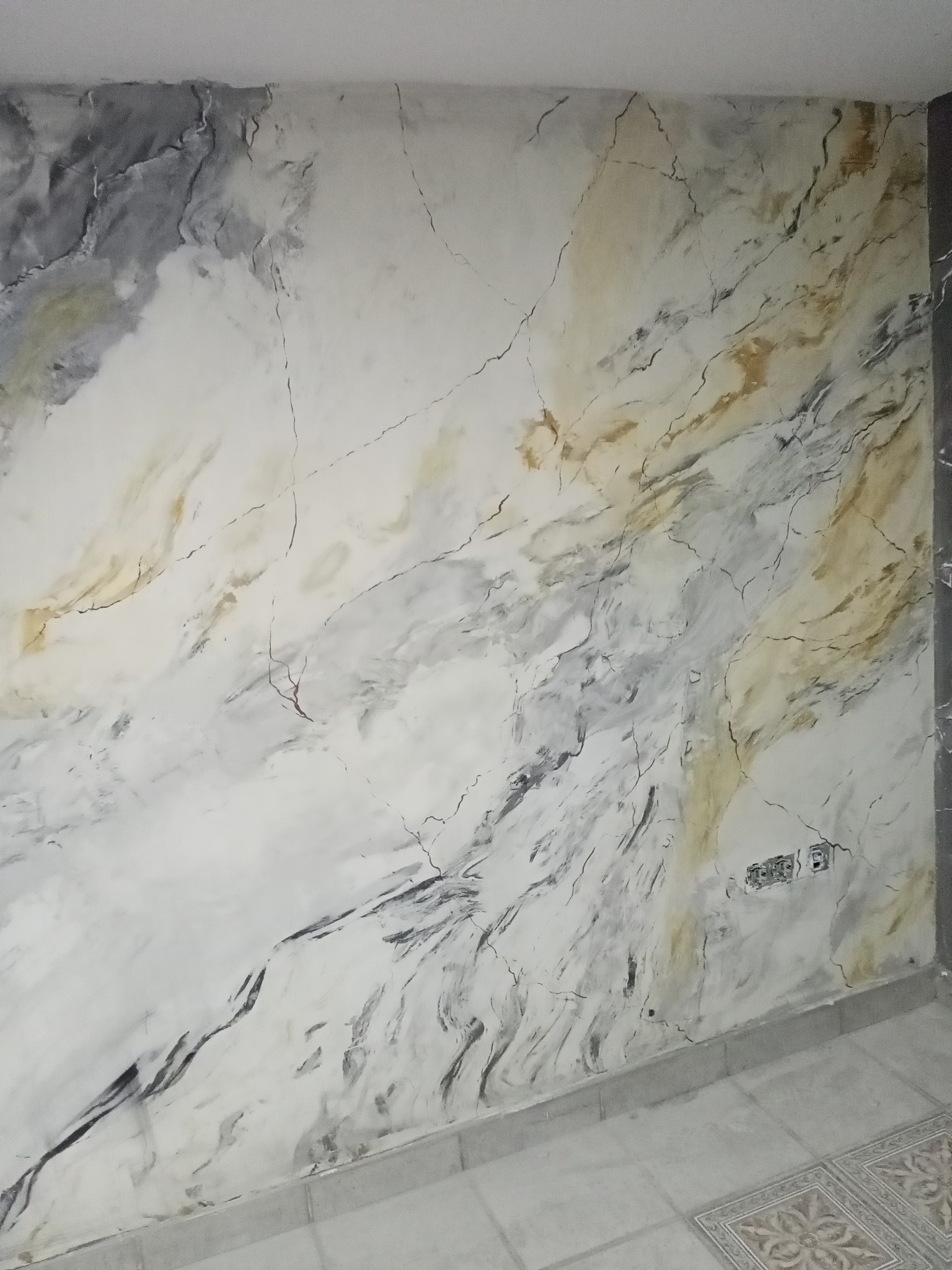 Ściana w pomieszczeniu wykończona dekoracyjną farbą imitującą marmur w odcieniach bieli, szarości i złota, z widocznymi drobnymi pęknięciami i gniazdkami elektrycznymi.