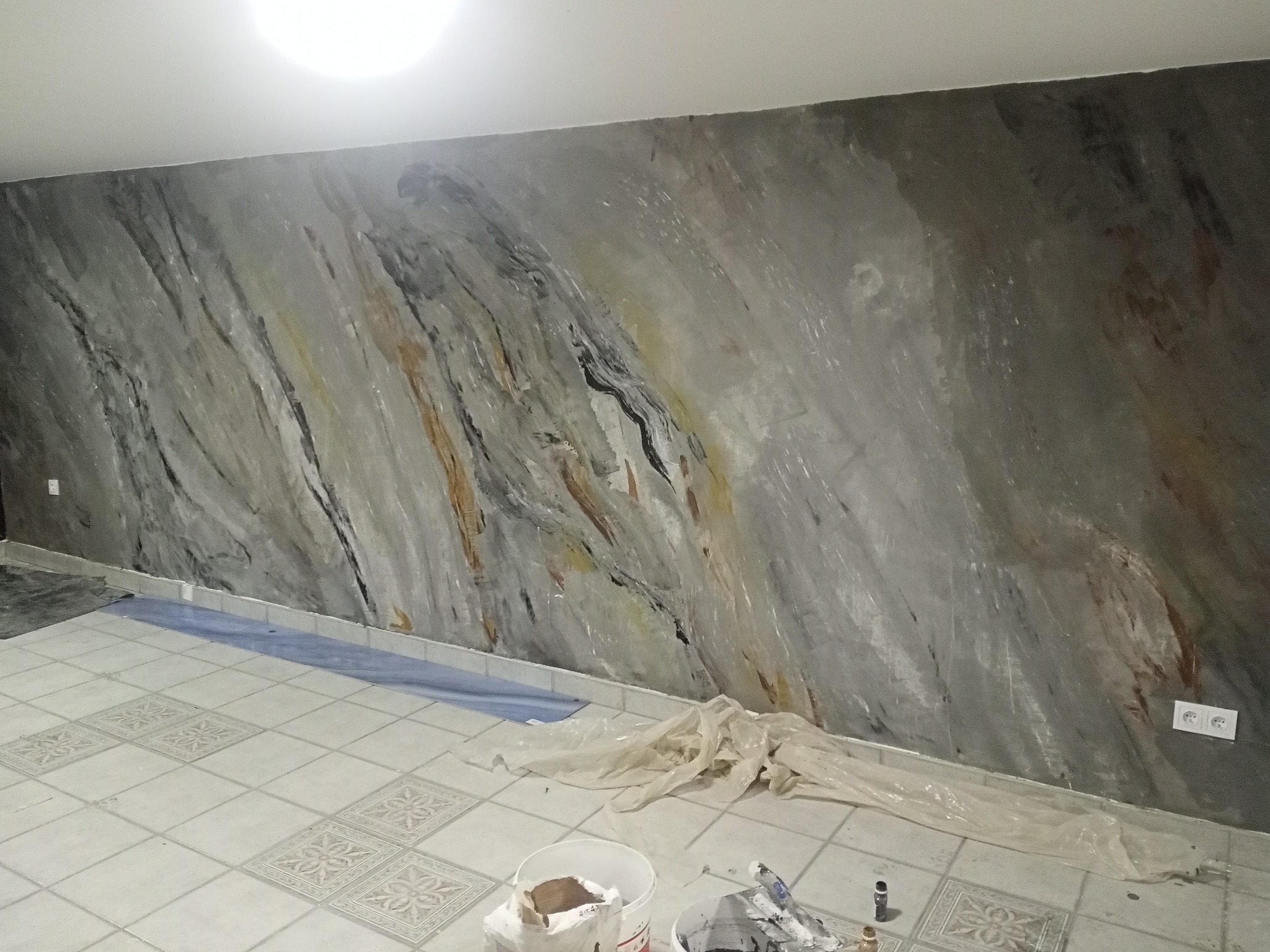 Ściana w pokoju z efektem marmuru, wykonana w odcieniach szarości, bieli i brązu. Na podłodze białe płytki z wzorem, widoczne wiaderka z farbą i folia ochronna.