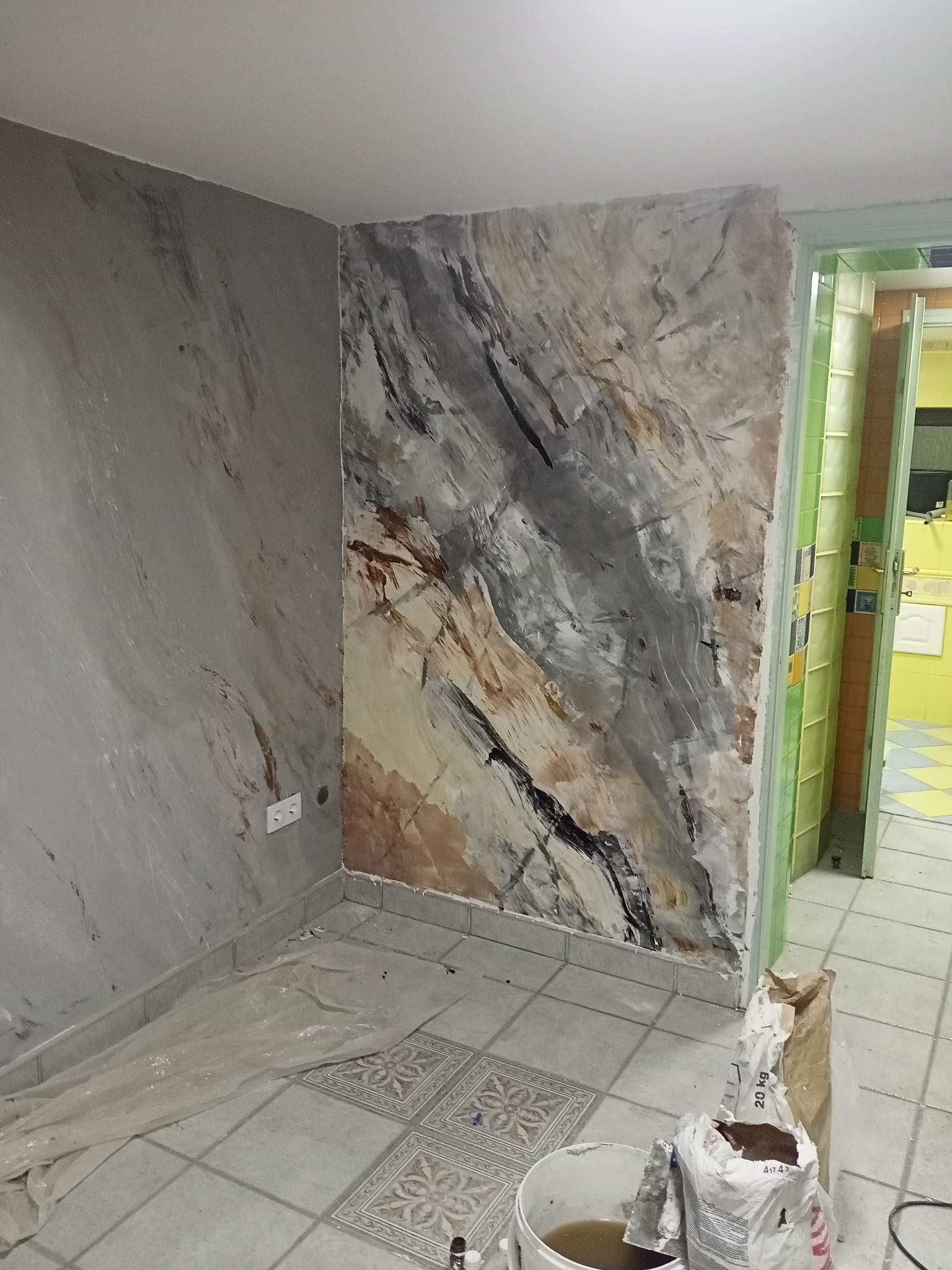 Ściany w trakcie dekoracyjnego wykończenia imitującego marmur w odcieniach szarości, beżu i czerni, widoczne materiały budowlane i wiadro z zaprawą, zabezpieczona folią podłoga w pomieszczeniu...