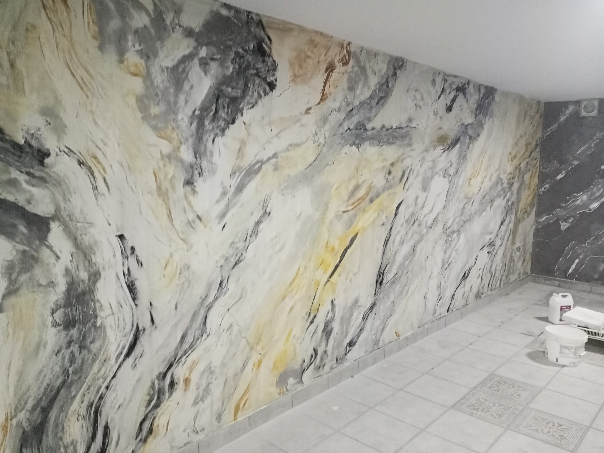 Ściana wykończona dekoracyjną techniką malarską imitującą marmur w odcieniach bieli, szarości i złota, z widocznymi wiaderkami farby i akcesoriami malarskimi na podłodze z płytek.