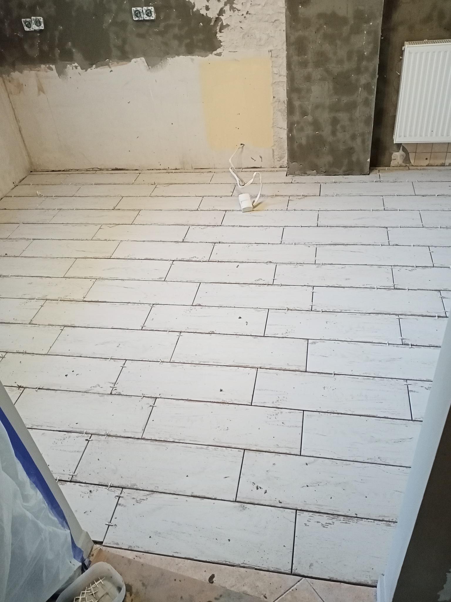 Ułożona jasna podłoga z płytek imitujących drewno w trakcie remontu, widoczne krzyżyki dystansowe, ściany częściowo przygotowane pod malowanie, grzejnik.