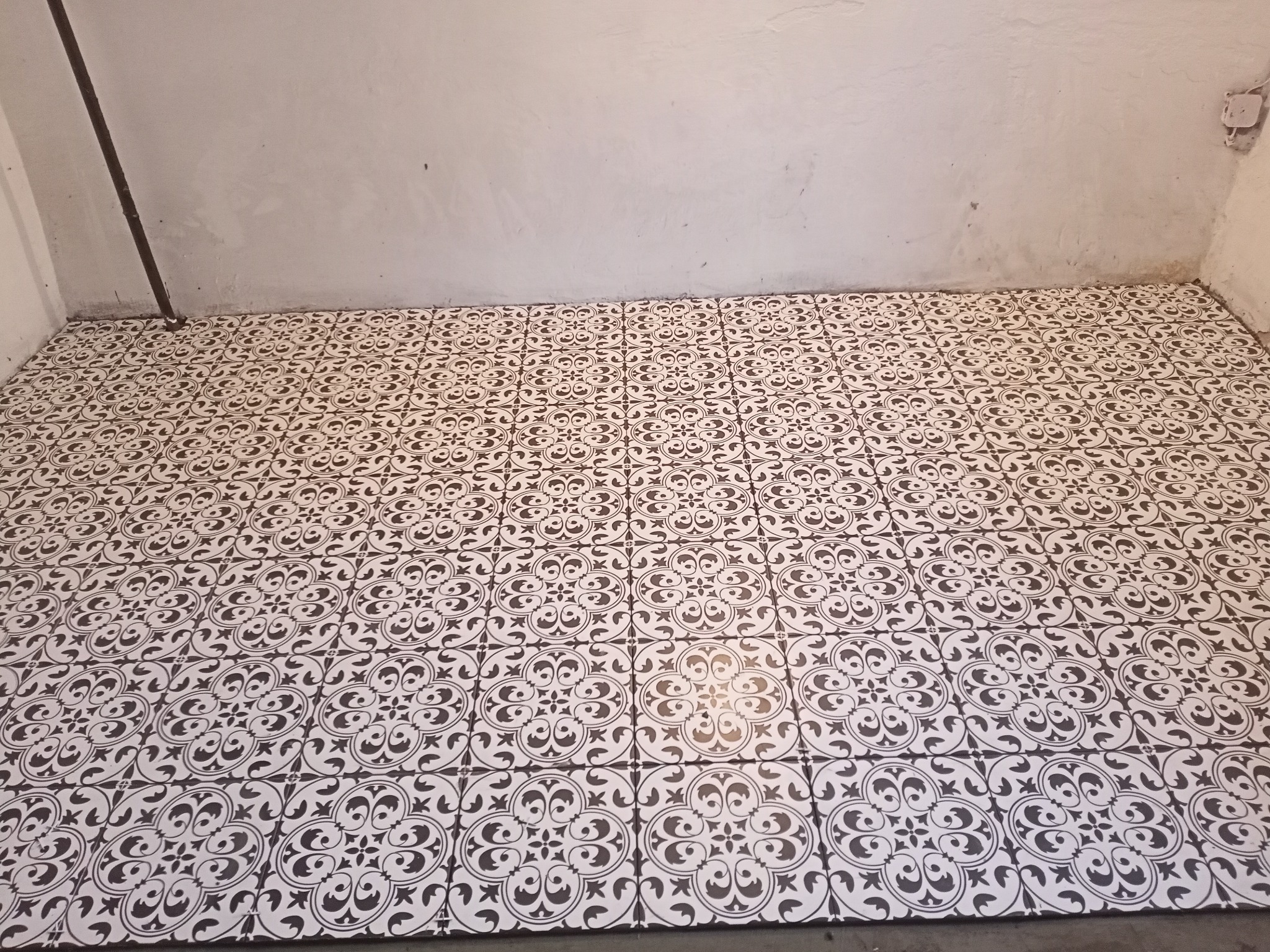 Ułożona podłoga z białych płytek z ciemnym, wzorzystym ornamentem w pomieszczeniu z surowymi, szarymi ścianami i widoczną instalacją elektryczną w rogu.