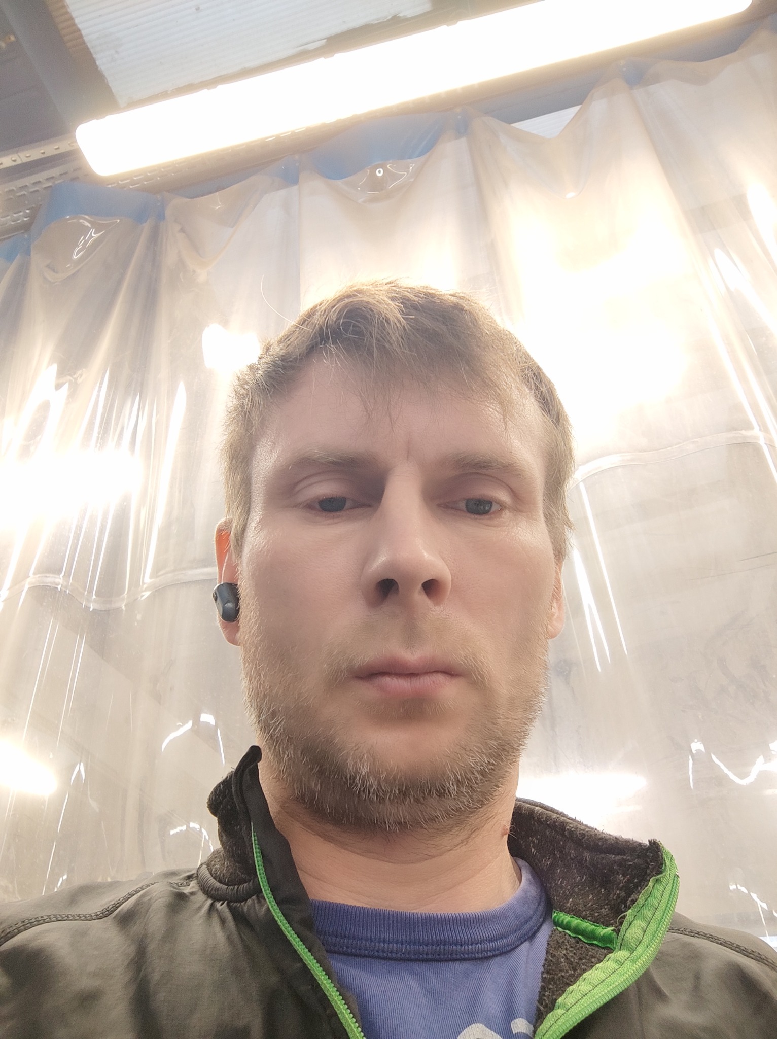 Autoportret elektryka z lampą LED i folią ochronną w tle, widoczne słuchawki bezprzewodowe w uchu.