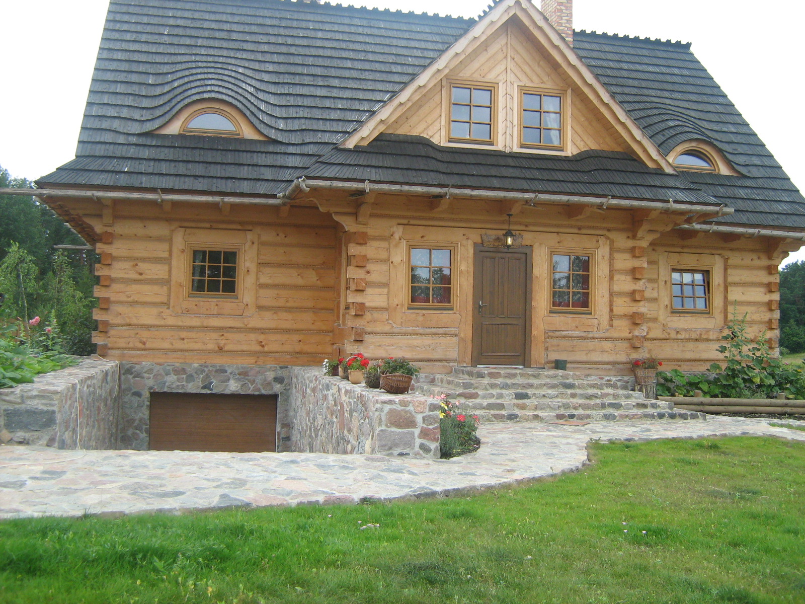 Drewniany dom z bali, z charakterystycznym dachem krytym gontem i kamiennym podjazdem do garażu, otoczony zielenią.