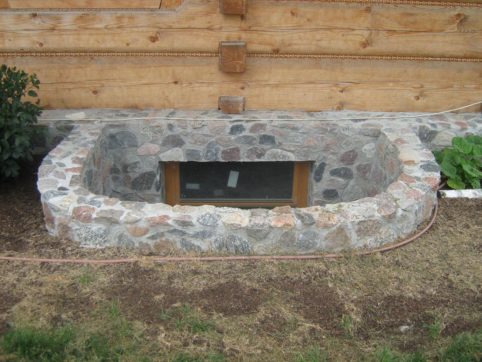 Kamienna opaska okienna piwniczna z drewnianą ramą, wkomponowana w elewację domu z bali; widoczny fragment trawnika i krzewów.