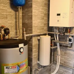 Wnętrze kotłowni z zamontowanym zbiornikiem buforowym, grzejnikiem i niebieskim zbiornikiem wyrównawczym. Widoczna instalacja hydrauliczna z rurami PCV na tle płytek ceramicznych.