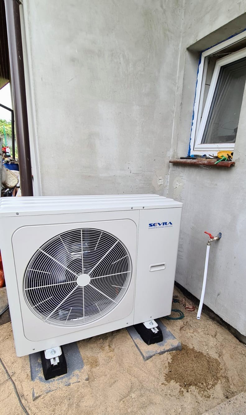 Zewnętrzna jednostka klimatyzacji marki Seyra zamontowana na betonowych podstawach przy ścianie budynku w Dębostrowie, widoczny zawór wodny i narzędzia na parapecie okna.