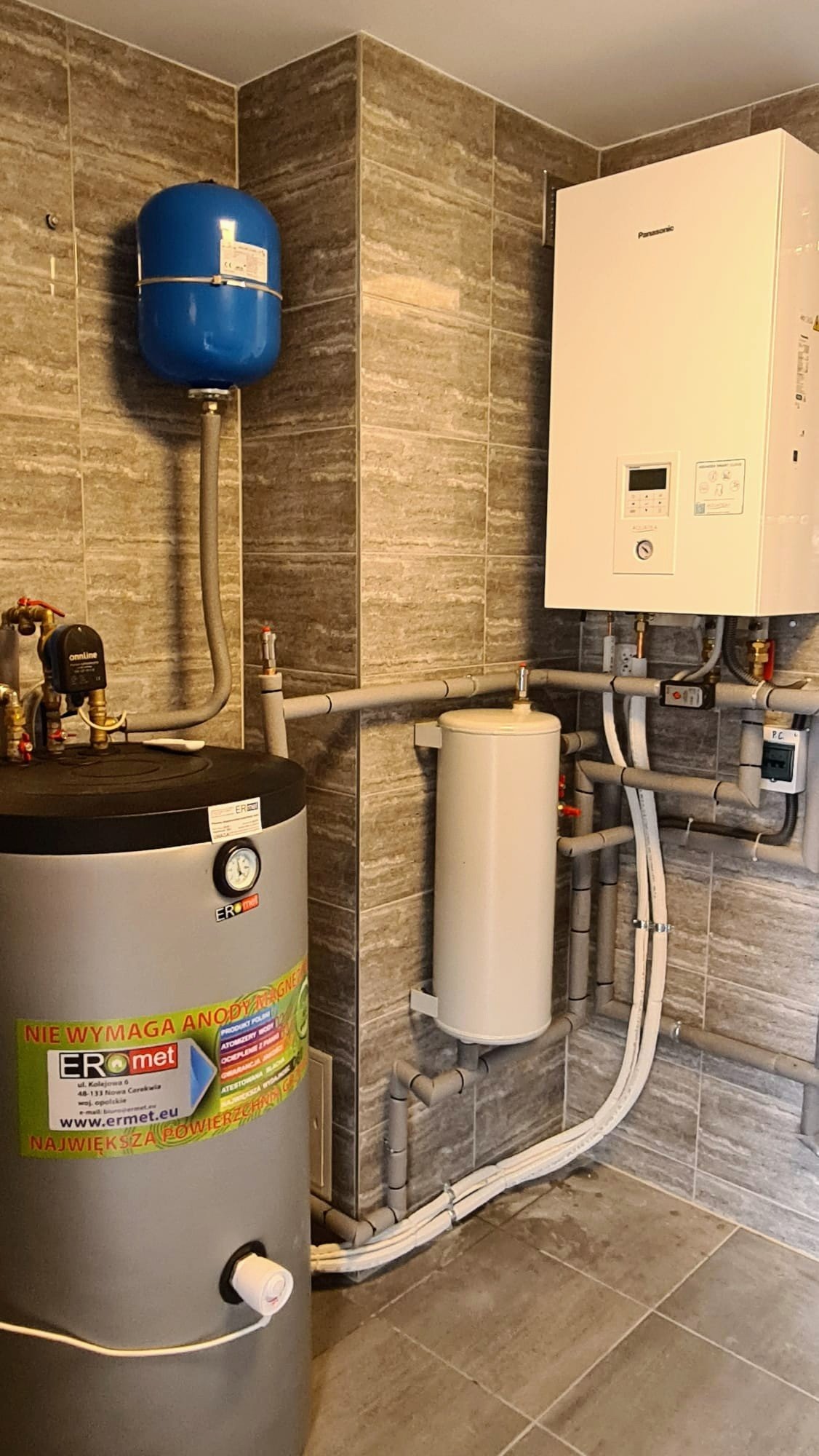 Wnętrze kotłowni z zamontowanym zbiornikiem buforowym, grzejnikiem i niebieskim zbiornikiem wyrównawczym. Widoczna instalacja hydrauliczna z rurami PCV na tle płytek ceramicznych.