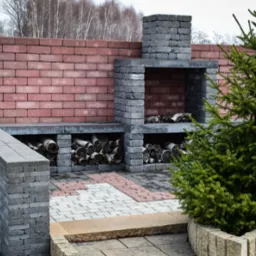 Murowany grill z szarej kostki betonowej z miejscem na drewno, obok mur z czerwonej cegły i mała choinka, ułożona kostka betonowa na ziemi.