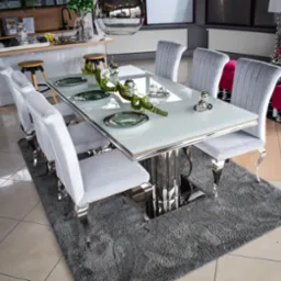 Elegancki stół jadalny z białym blatem i chromowanymi nogami, otoczony sześcioma krzesłami z szarym, welurowym obiciem i chromowanymi nogami, na szarym, puszystym dywanie.