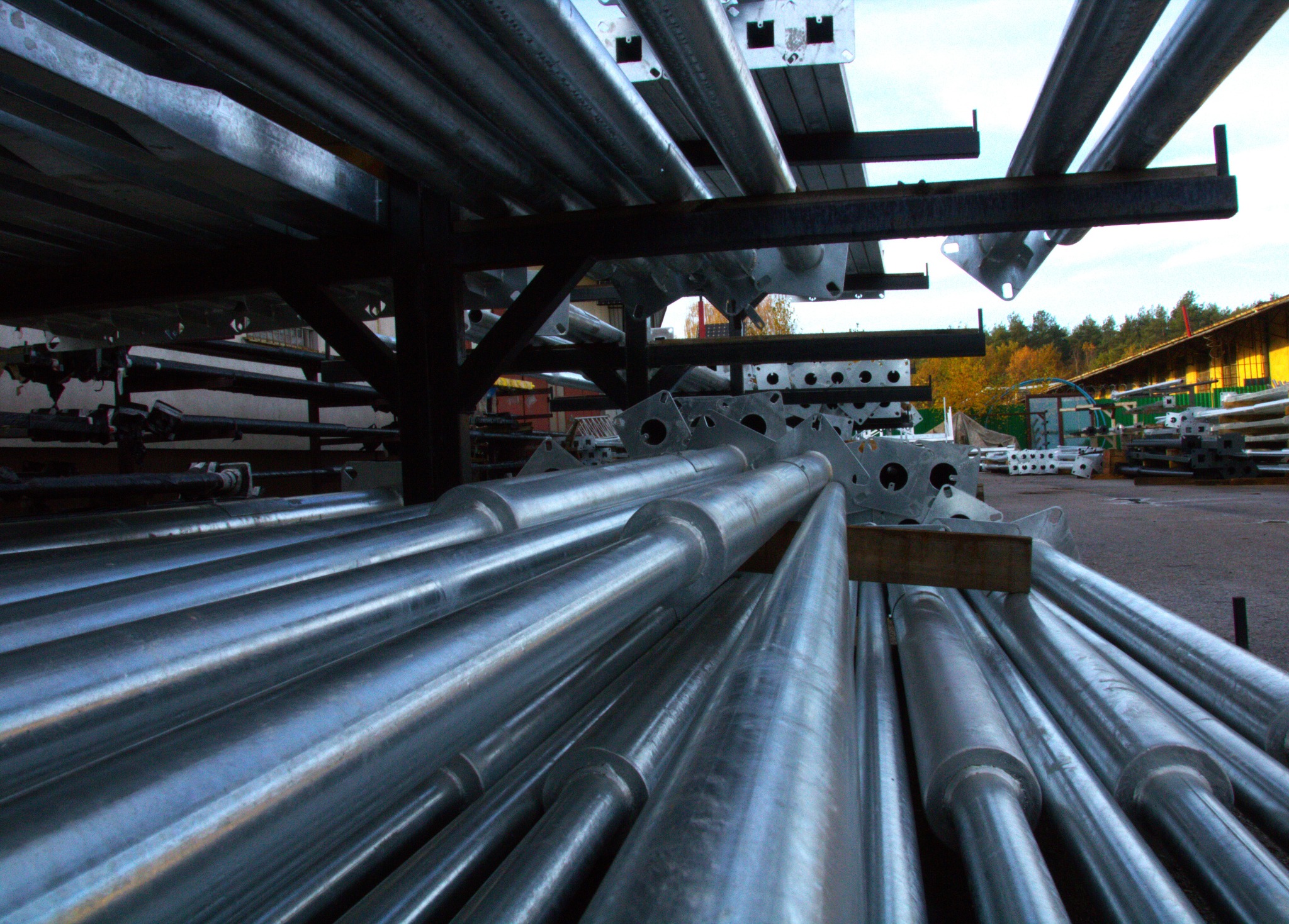 Magazyn prefabrykatów betonowych: stosy metalowych rur i elementów konstrukcyjnych na zewnątrz, widoczne elementy z otworami i mocowaniami, w tle budynek magazynowy i drzewa.