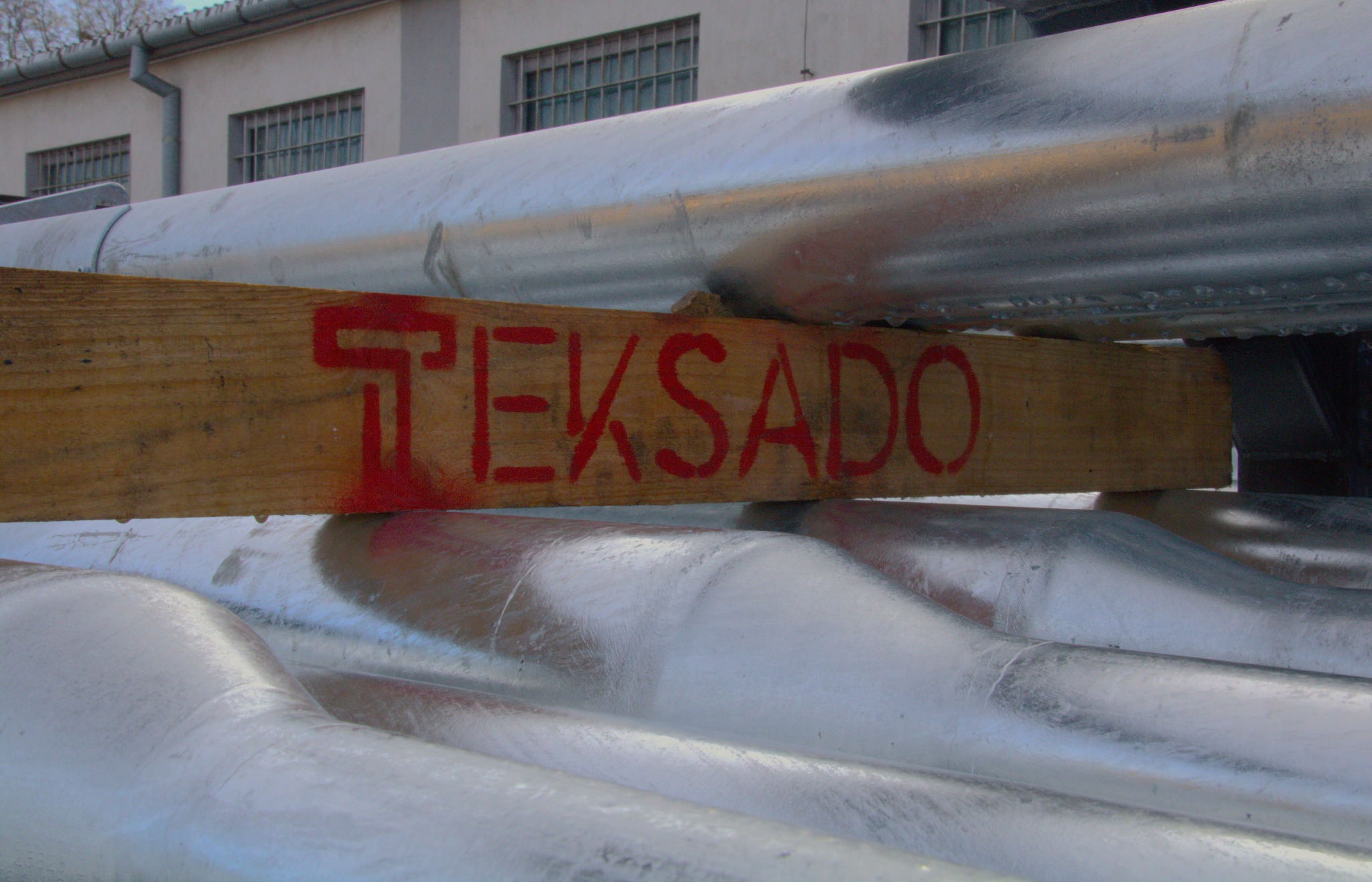 Stos prefabrykowanych elementów betonowych, zabezpieczonych drewnianą belką z czerwonym napisem TEKSADO, na tle budynku.