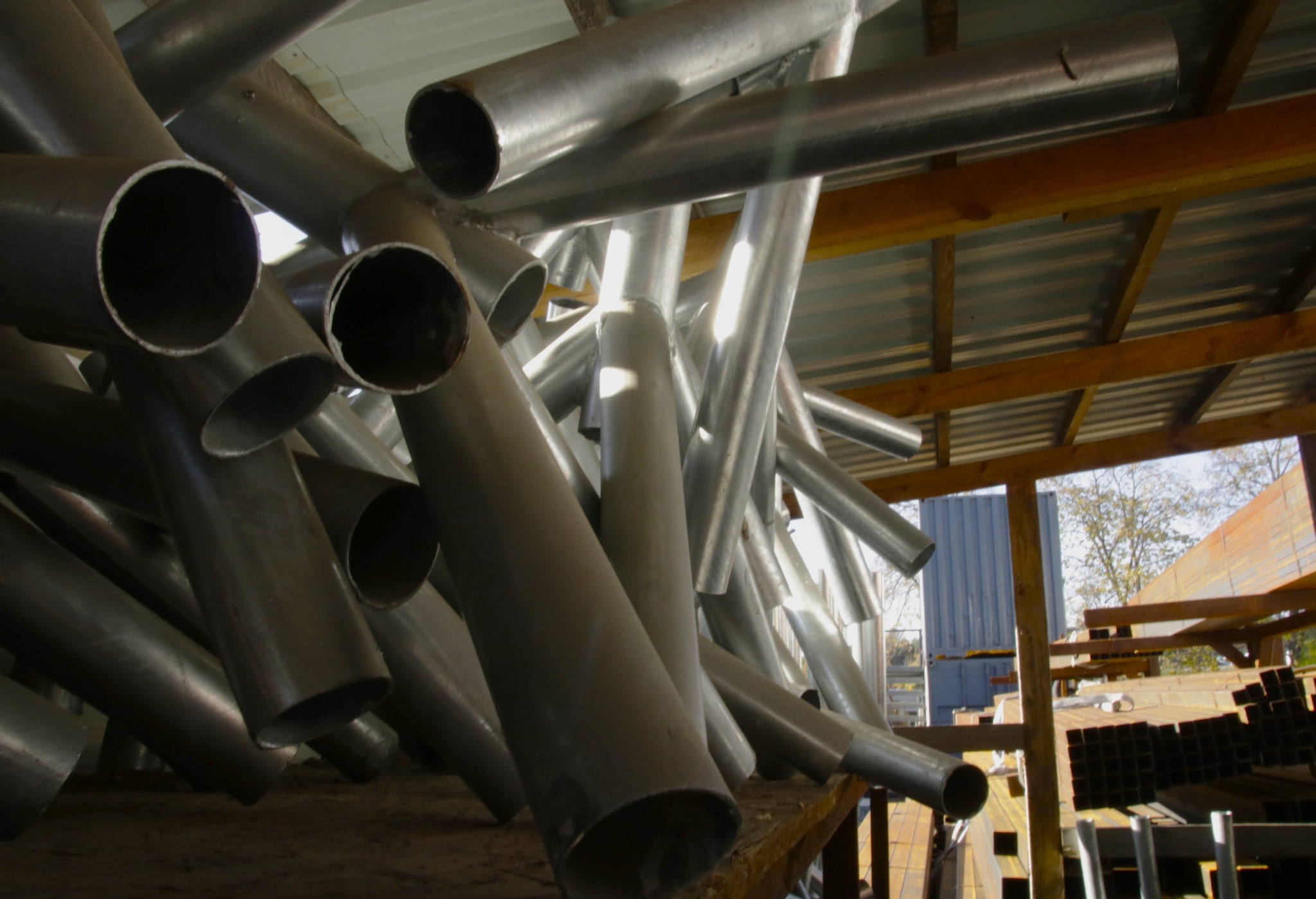 Stalowe rury o różnych średnicach i długościach, ułożone chaotycznie pod zadaszeniem, z widocznymi elementami konstrukcyjnymi dachu i fragmentem otoczenia zewnętrznego w tle.