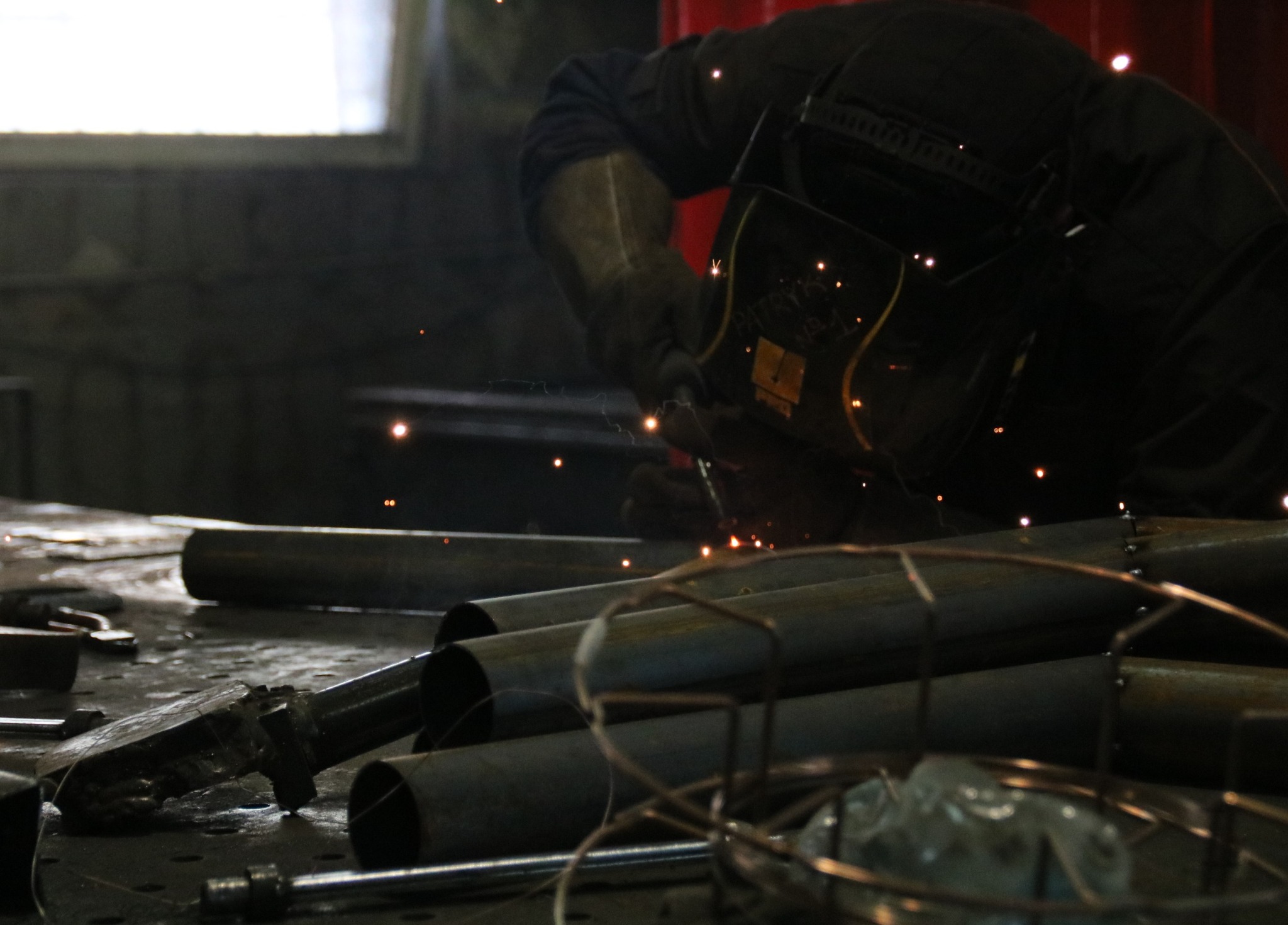Spawanie rur na warsztacie: spawacz w masce ochronnej łączy metalowe elementy, widoczne iskry i narzędzia na stole warsztatowym.