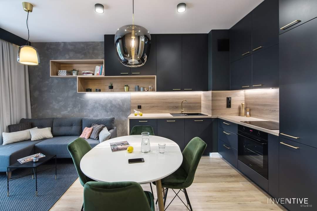 Nowoczesna kuchnia w odcieniach granatu z drewnianymi akcentami, oświetleniem LED pod szafkami, złotymi uchwytami i okrągłym stołem z zielonymi krzesłami w tle salonu z szarą sofą.