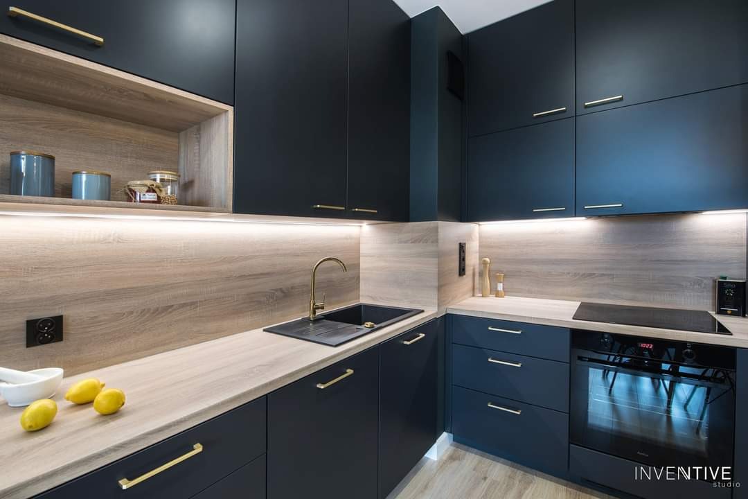 Nowoczesna, narożna kuchnia z ciemnymi, granatowymi szafkami, złotymi uchwytami i drewnopodobnym blatem, oświetlona listwą LED, z czarnym zlewem i złotą baterią oraz trzema cytrynami w pobliżu.