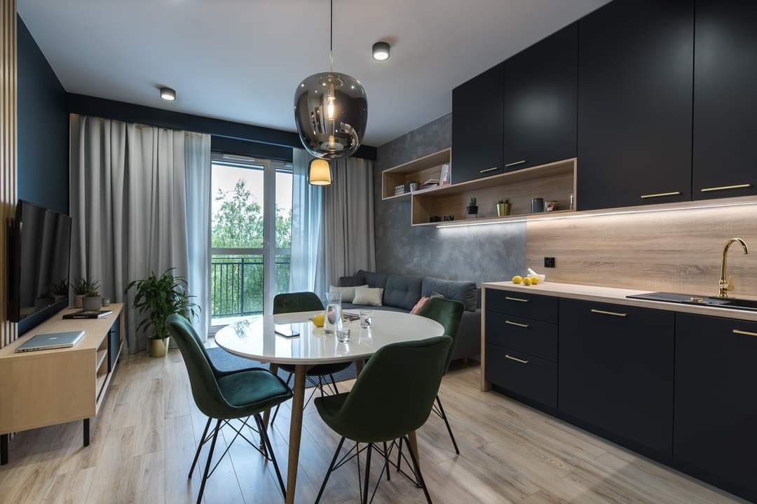 Nowoczesna kuchnia w odcieniach granatu i drewna, z okrągłym stołem i zielonymi krzesłami, oświetlona wiszącą lampą w kształcie kuli, widok na salon z szarą sofą w tle.