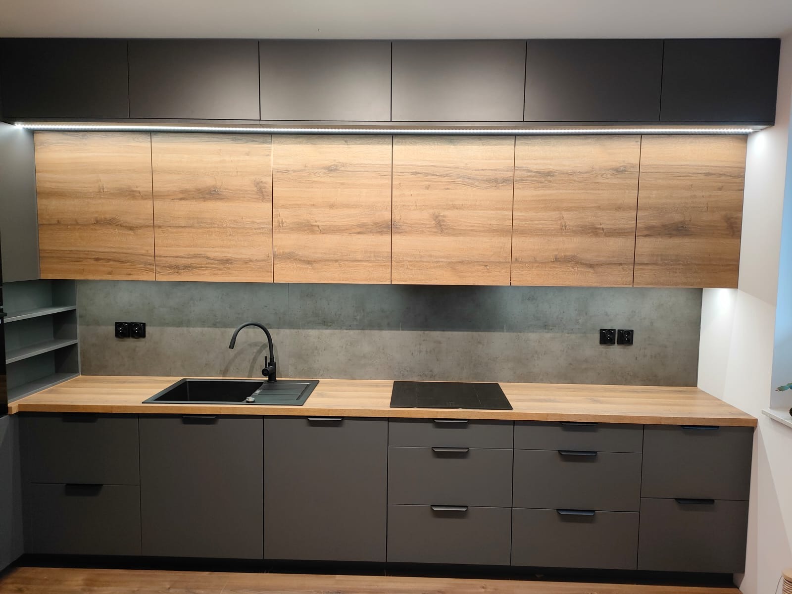 Nowoczesna kuchnia z szarymi szafkami dolnymi i drewnopodobnymi szafkami górnymi, oświetlenie LED pod szafkami górnymi, czarny zlew i płyta indukcyjna.