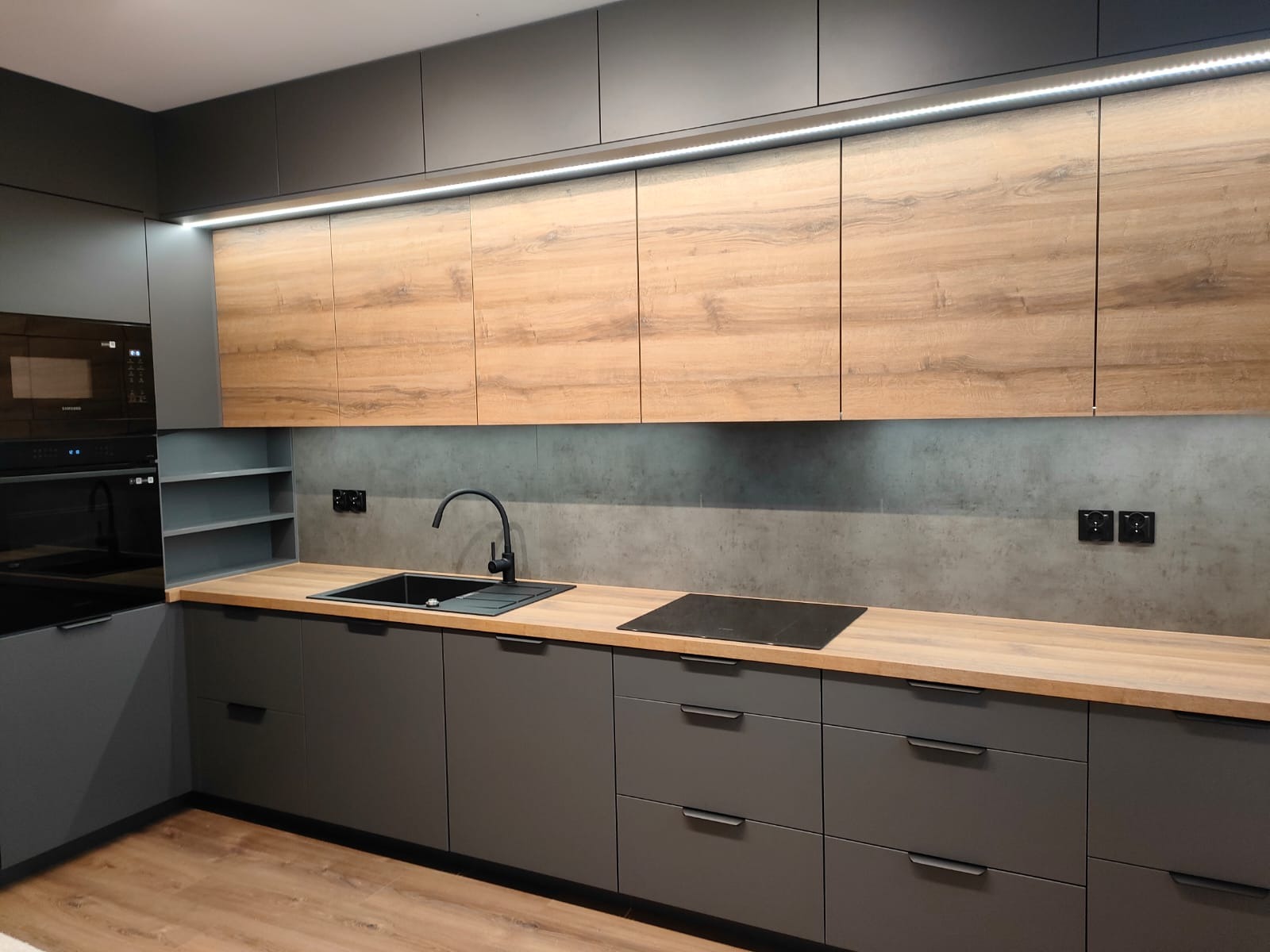 Nowoczesna kuchnia w odcieniach szarości i drewna, z podświetleniem LED, czarnym zlewem i płytą indukcyjną, widok na drewnianą podłogę.