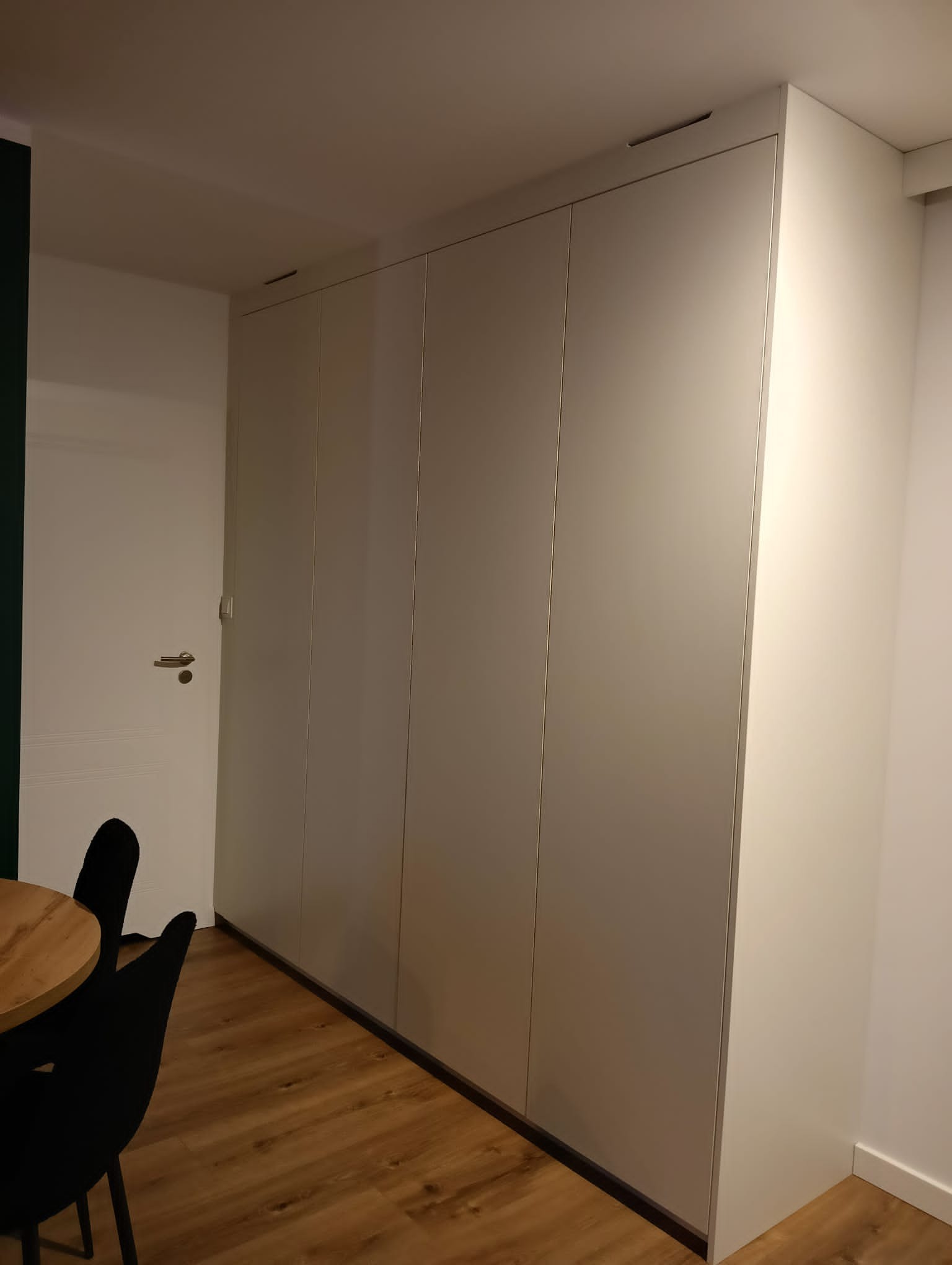 Biała szafa wnękowa z czterema frontami, wbudowana od podłogi do sufitu, widoczna w rogu pokoju z drewnianą podłogą i fragmentem stołu z krzesłami.