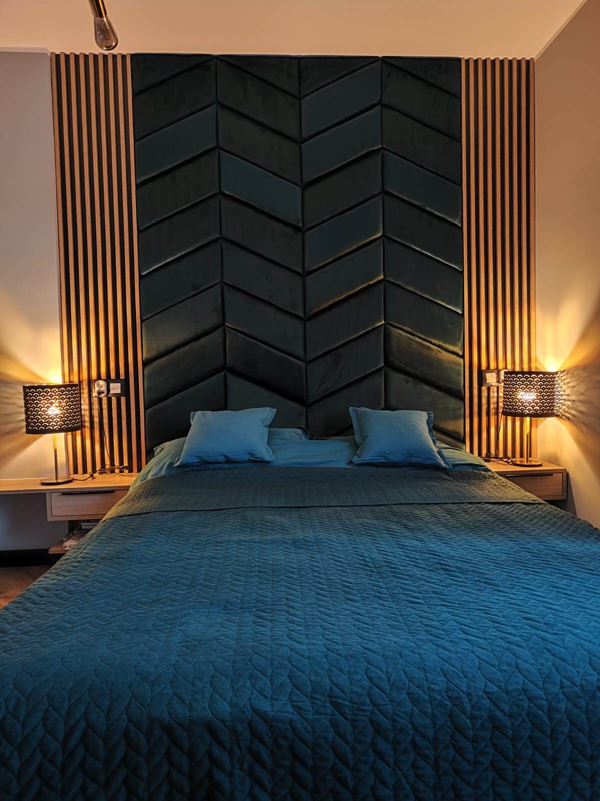 Sypialnia z ciemnozielonym zagłówkiem w jodełkę, drewnianymi lamelami po bokach i szafkami nocnymi z lampkami o ażurowych abażurach.