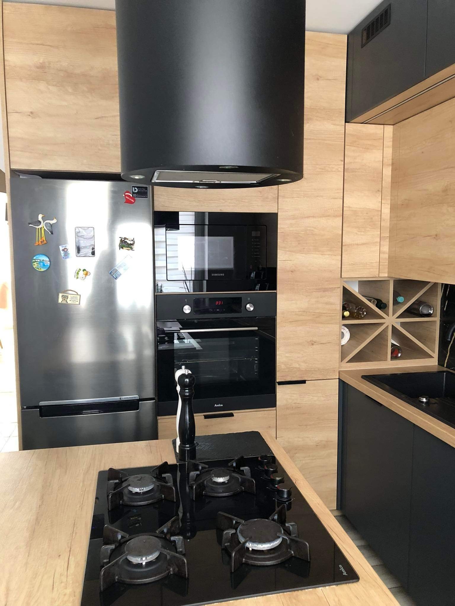 Nowoczesna kuchnia z frontami w kolorze jasnego drewna, czarnym okapem i płytą gazową, lodówka ozdobiona magnesami.