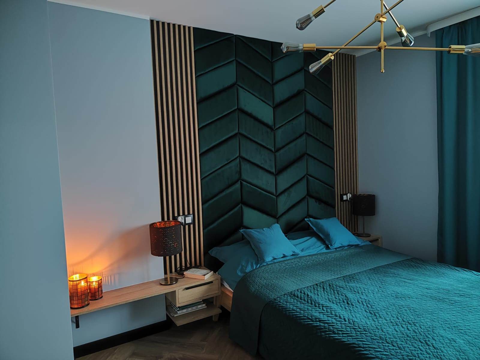 Sypialnia z łóżkiem z zielonym zagłówkiem w jodełkę, drewnianymi panelami, lampkami nocnymi i złotym żyrandolem.