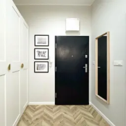 Nowoczesny przedpokój z czarnymi drzwiami wejściowymi, podłogą w jodełkę, białymi szafami w zabudowie i lustrem w drewnianej ramie.